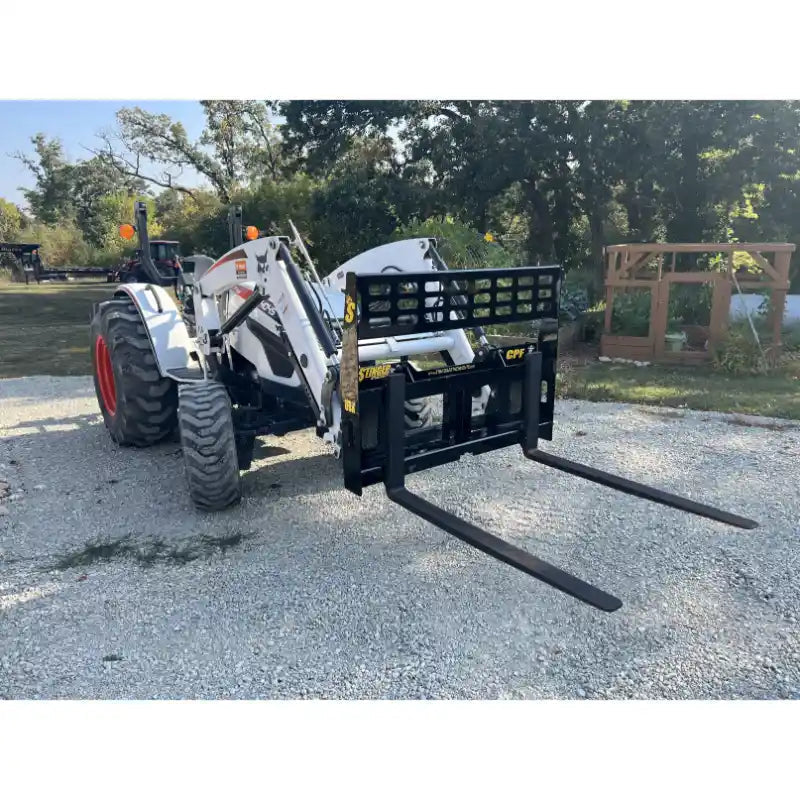 White tractor with front hydraulic loader and black steel pallet fork attachment on gravel surface outdoors