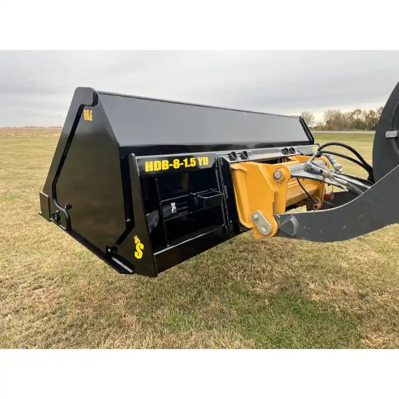 Close-up of black steel hydraulic dump bucket attachment connected to yellow tractor loader arm over grass field