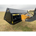 Black steel hydraulic dump bucket attachment mounted on loader arm with yellow linkage over grassy field under cloudy sky