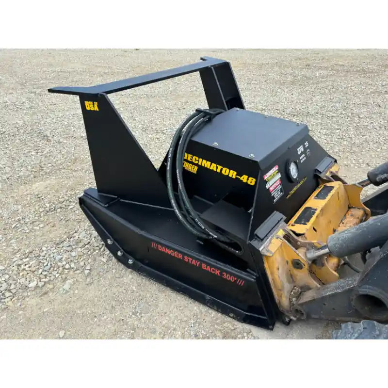 Black steel hydraulic mulcher attachment with hoses and warning labels connected to skid steer loader on gravel surface