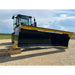 Large black and yellow skid steer snow pusher attachment mounted on loader on gravel surface with field background