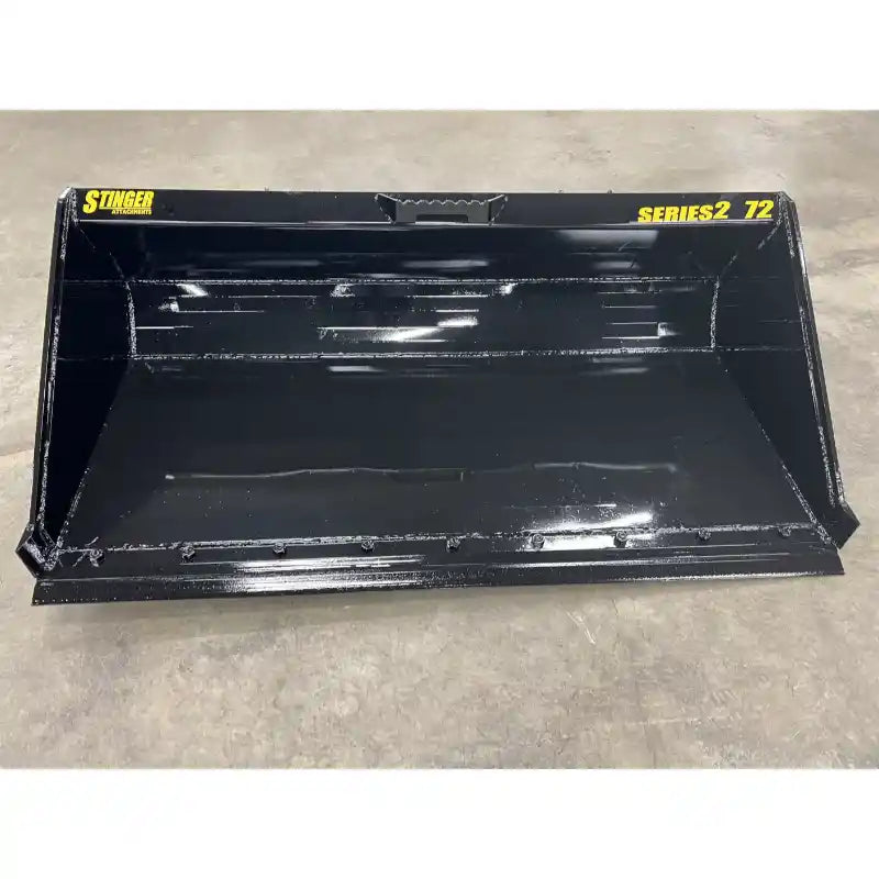 Black steel skid steer material bucket with smooth interior and yellow Stinger Series 2 72 markings on concrete floor