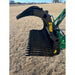 Black steel rock grapple attachment with serrated upper jaw and spaced tines connected to green loader arm over dry grass