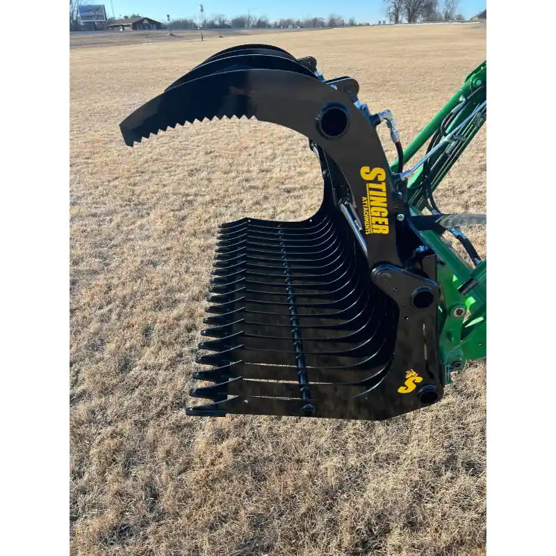 Black steel rock grapple attachment with serrated upper jaw and spaced tines connected to green loader arm over dry grass