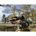 Yellow skid steer loader equipped with black grapple attachment lifting large tree logs outdoors on grass and dirt