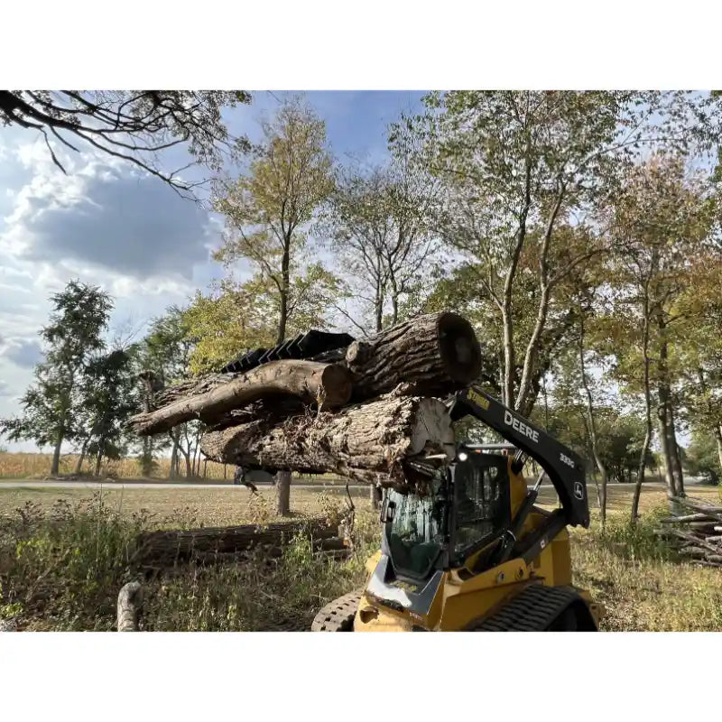 Yellow skid steer loader equipped with black grapple attachment lifting large tree logs outdoors on grass and dirt