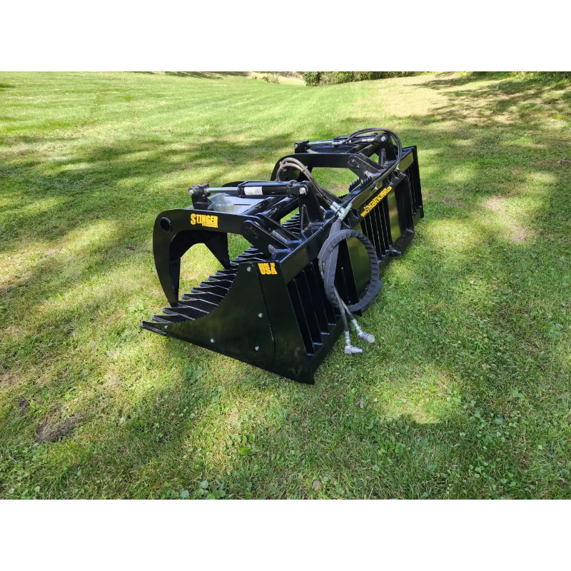 Black metal skid steer grapple attachment with spaced tines, hydraulic arms, hose connections, and reinforced side plates on grass