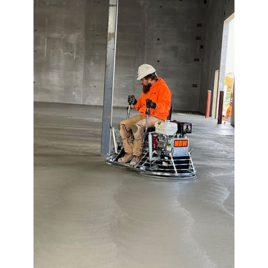 Operator seated on a ride-on power trowel smoothing a large concrete floor inside a building under construction