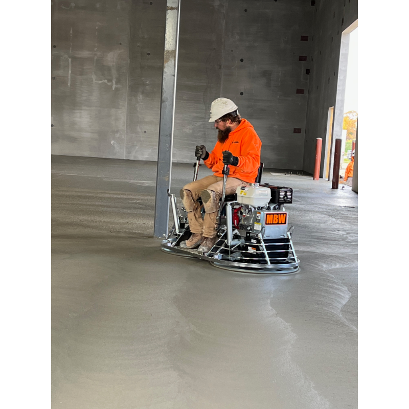 Operator seated on a ride-on power trowel smoothing a large concrete floor inside a building under construction