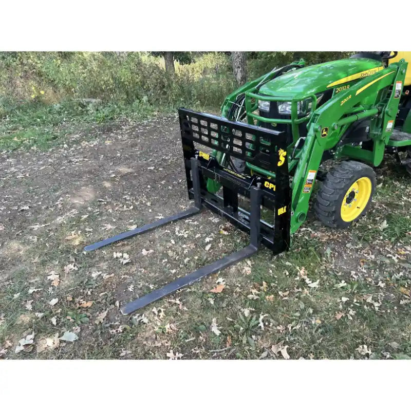 Green tractor with black steel pallet fork attachment featuring open grid backrest and extended fork tines on grass and dirt ground