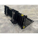 Black steel skid steer rock bucket attachment with wide spaced flat tines and reinforced mounting points on concrete floor
