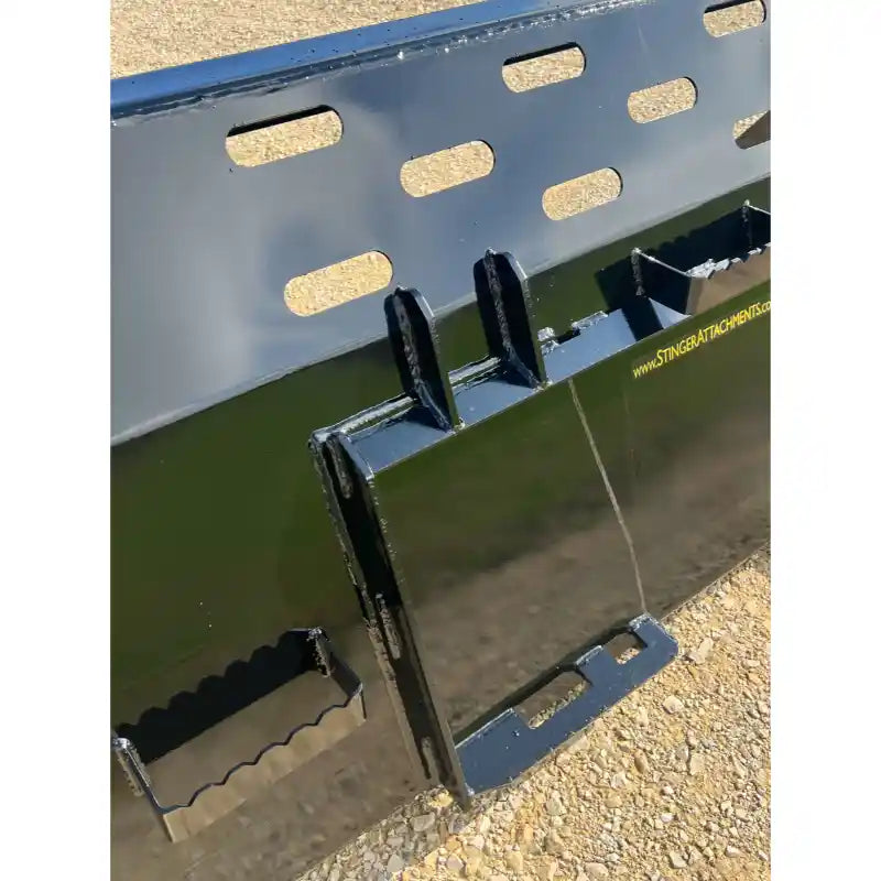 Close-up of black skid steer snow bucket attachment with mounting brackets and rectangular ventilation slots on gravel surface