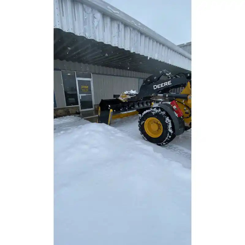 Yellow skid steer loader with DEERE arm lifting a black steel snow pusher attachment loaded with snow by a metal building