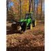 Green compact tractor with enclosed cab and front loader bucket carrying large rock in wooded area with fallen leaves
