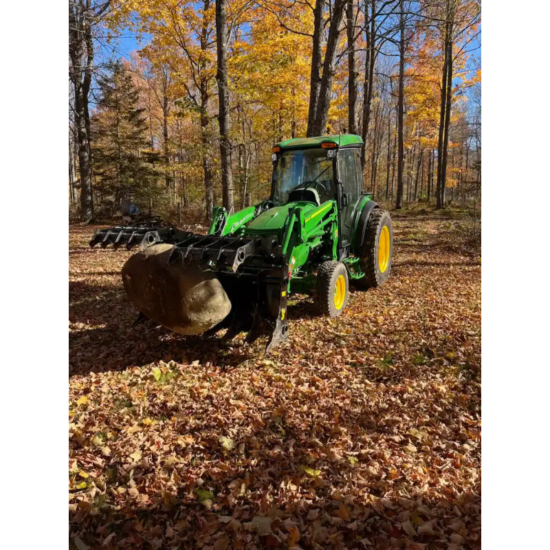 Green compact tractor with enclosed cab and front loader bucket carrying large rock in wooded area with fallen leaves