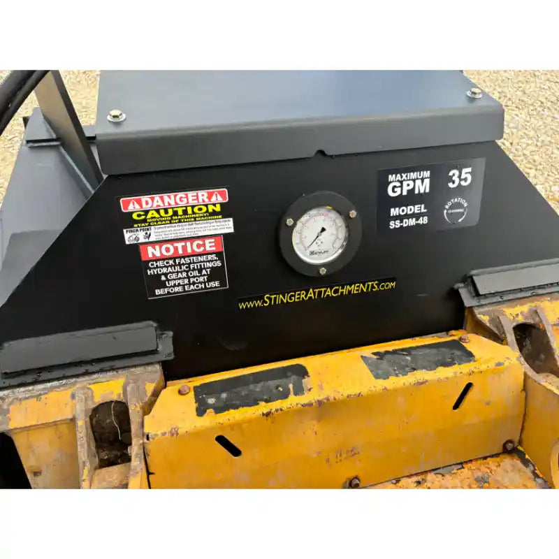 Close-up of black metal equipment panel with warning labels, pressure gauge, and yellow skid steer mounting bracket