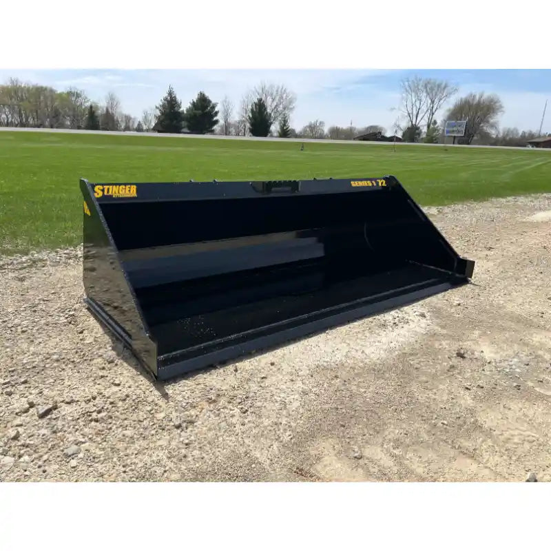 Black steel skid steer material bucket with smooth interior and yellow "Stinger Series 1 72" lettering on gravel and grass background