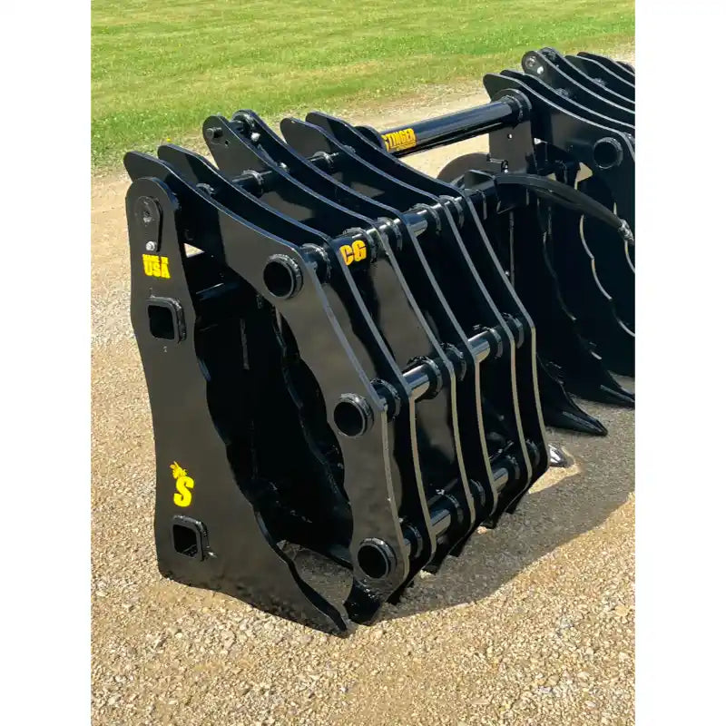 Black steel clam grapple attachment with multiple curved tines and hydraulic linkage resting on gravel outdoors