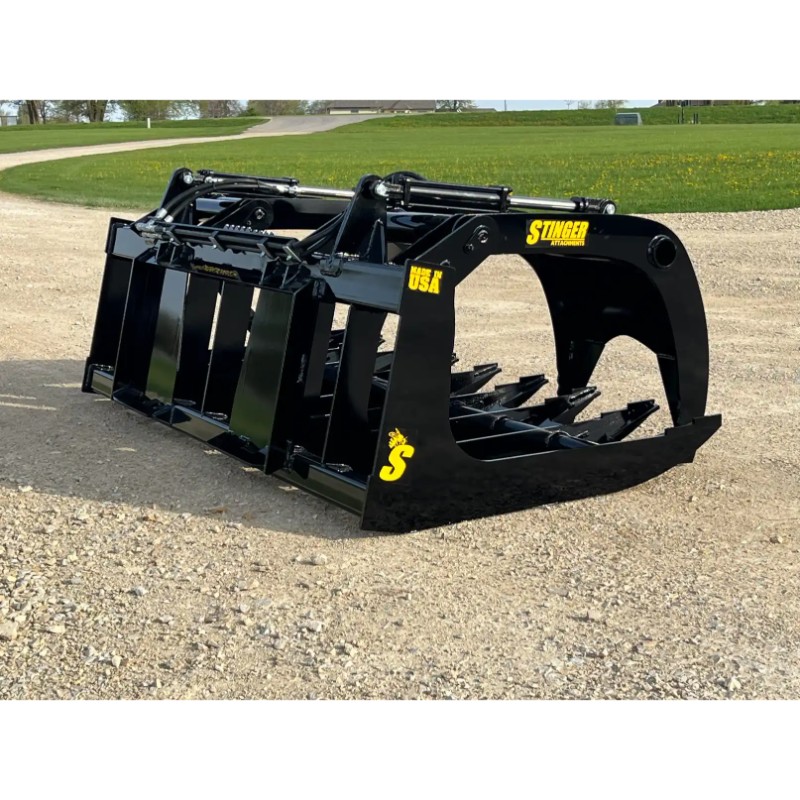 Black skid steer root grapple attachment with wide curved tines, hydraulic cylinders, and reinforced steel frame on gravel surface