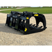 Black metal skid steer root grapple attachment with curved tines, hydraulic cylinders, and reinforced frame on gravel surface