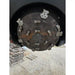 Round metal disc with multiple welded cutting teeth and mounting bolts resting above gravel and wooden pallets
