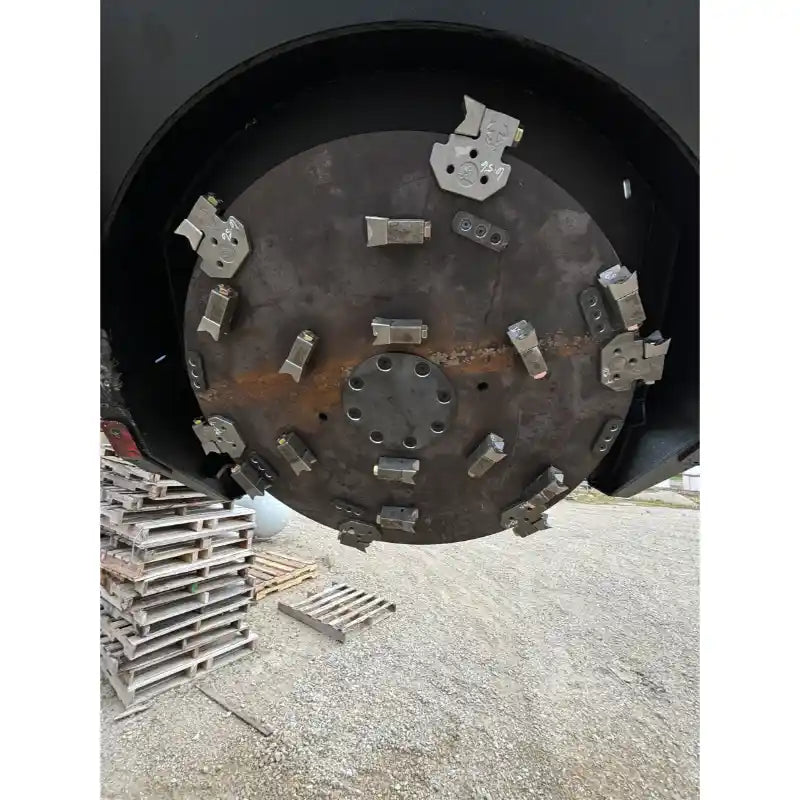 Round metal disc with multiple welded cutting teeth and mounting bolts resting above gravel and wooden pallets