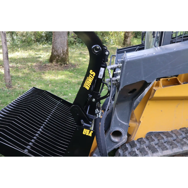 Side view of black metal skid steer rock grapple attachment with hydraulic hoses and yellow lettering on loader arm outdoors