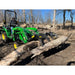 Green tractor with black hydraulic rock grapple attachment lifting large log in leafless wooded area on dirt ground