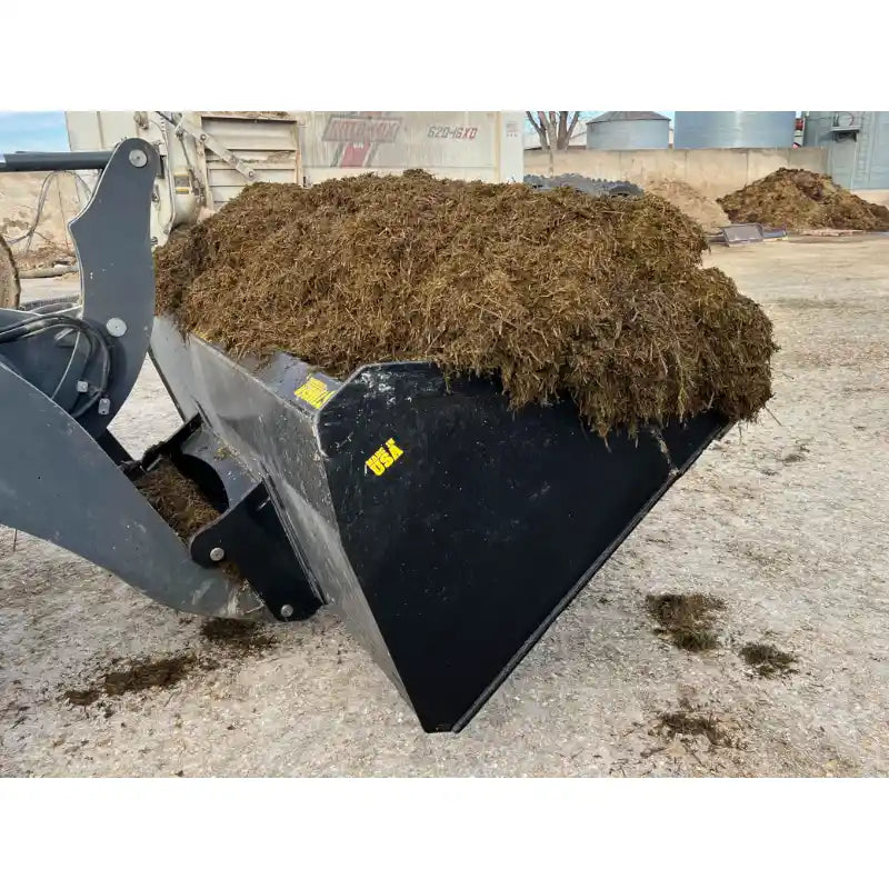 Black skid steer bucket attachment loaded with loose organic material at a gravel and concrete industrial yard