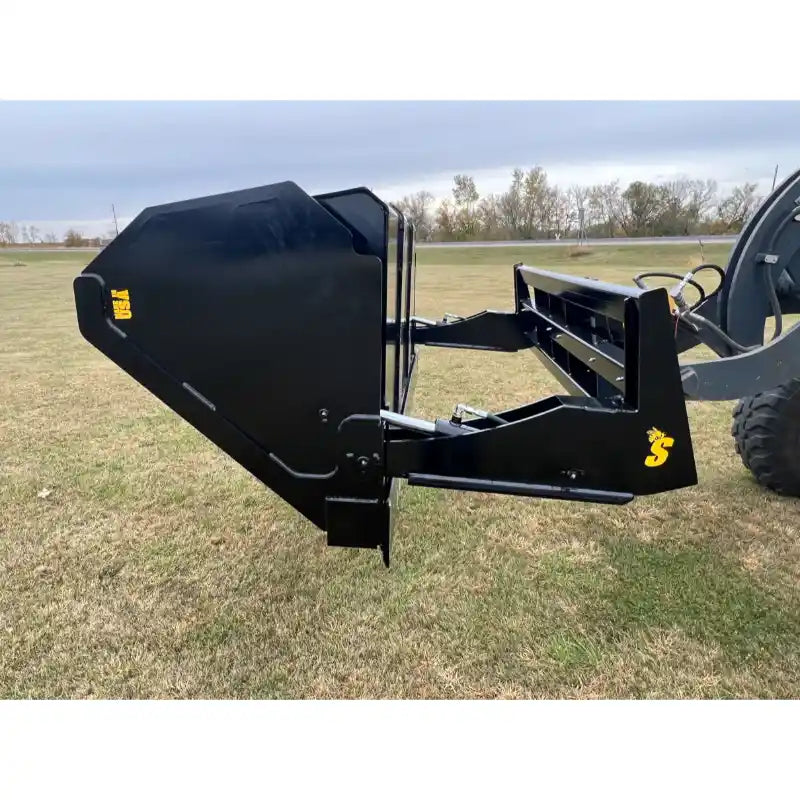 Side view of black steel hydraulic dump bucket attachment mounted on front loader arm over grass field