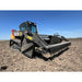 Low-angle front view of tracked skid steer with metal rock bucket attachment on dry tilled soil under clear sky
