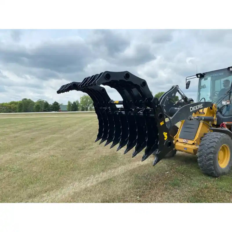 Yellow John Deere skid steer loader with large black multi-tine grapple attachment raised over dry grass field under cloudy sky