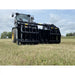 John Deere tractor with large black steel clam-style grapple attachment lowered over dry grass field under cloudy sky