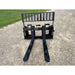 Black steel pallet fork attachment with perforated vertical backrest and two long flat tines on gravel surface