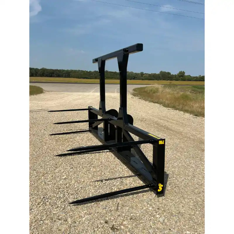 Black steel loader attachment with five long pointed bale spears and reinforced upright supports on gravel surface outdoors