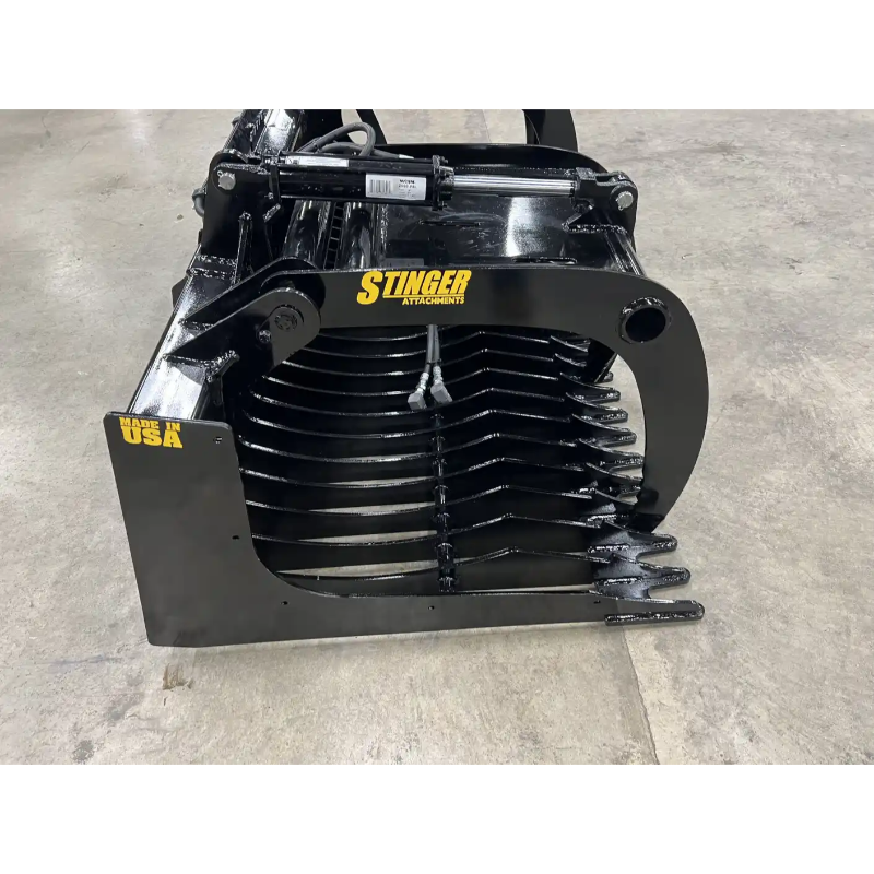 Black painted steel skid steer grapple attachment with curved tines, hydraulic cylinder, and reinforced side plate on concrete floor