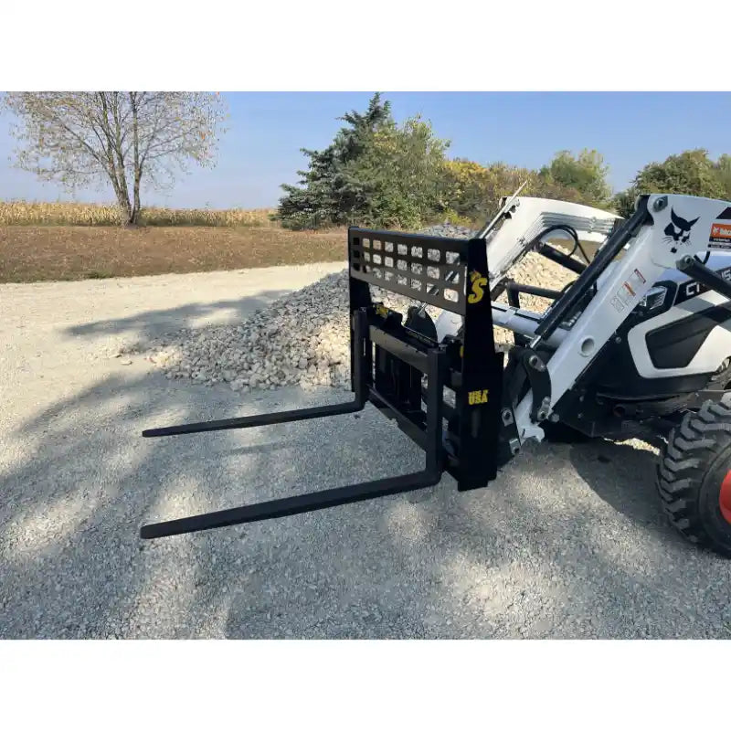 Compact black steel pallet fork attachment with perforated backrest mounted on white skid steer outdoors on gravel surface