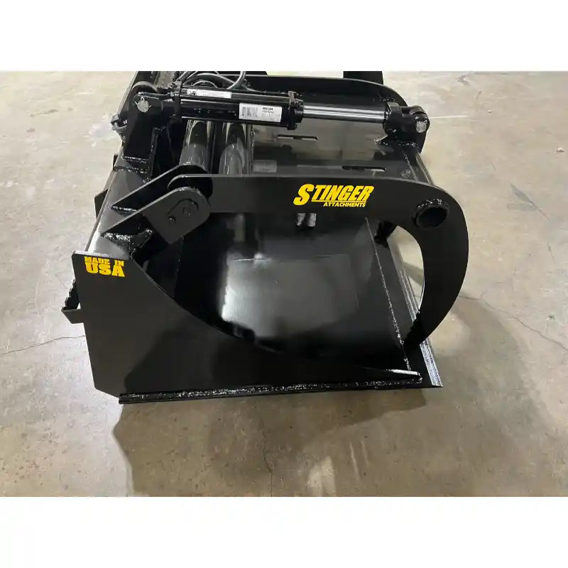Black skid steer grapple bucket attachment with hydraulic cylinder and clamp arm on concrete floor indoors