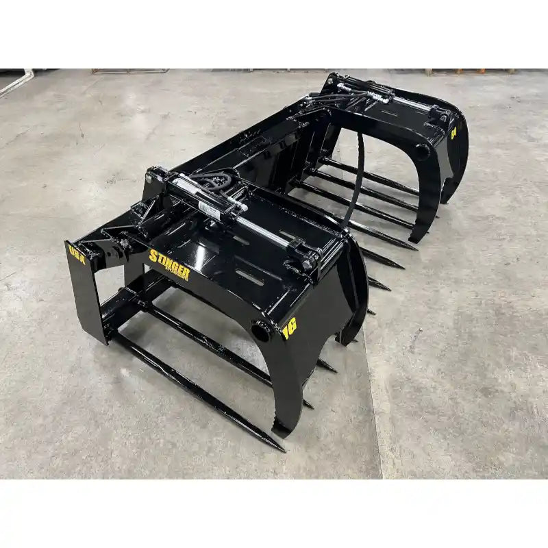 Pair of black steel grapple attachments with curved tines and hydraulic cylinders on concrete floor indoors