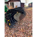 Black steel grapple attachment on green loader lifting large rock among fallen leaves outdoors near wooden buildings