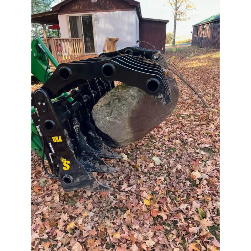 Black steel grapple attachment on green loader lifting large rock among fallen leaves outdoors near wooden buildings
