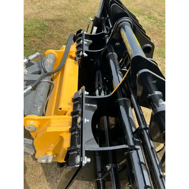 Close-up view of black steel hydraulic grapple attachment mounted on yellow loader arm over grassy terrain