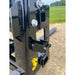 Close-up of black steel skid steer attachment plate with serrated locking latch and two mounting slots on gravel surface