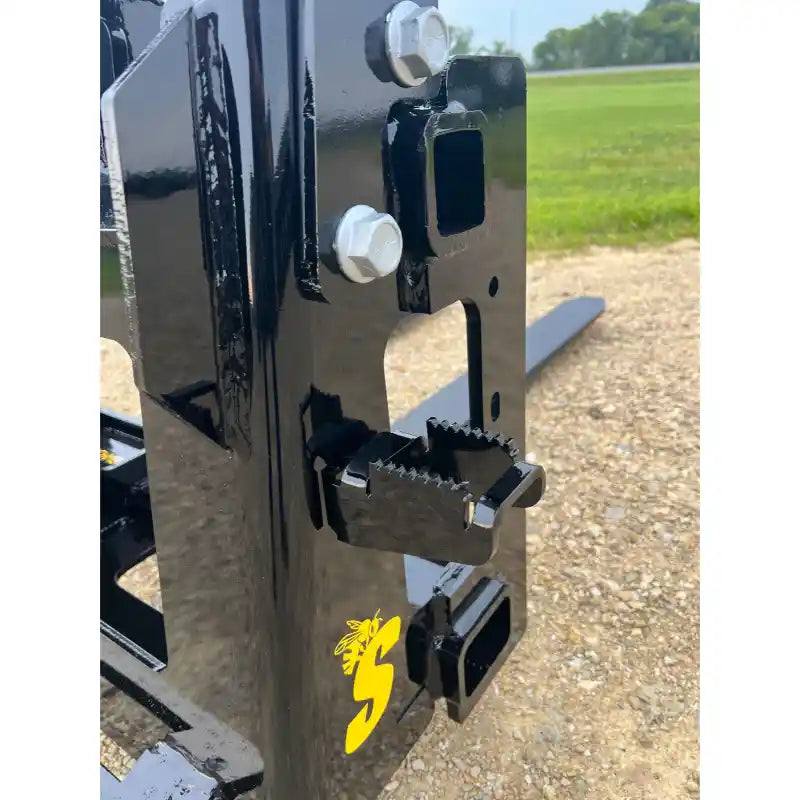 Close-up of black steel skid steer attachment plate with serrated locking latch and two mounting slots on gravel surface