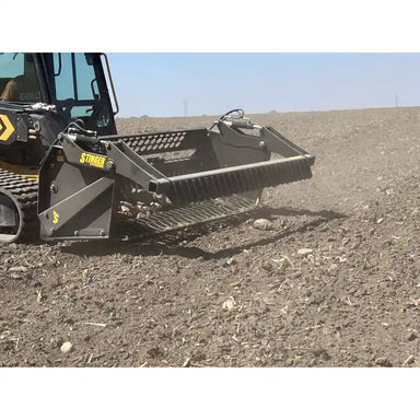 Side view of skid steer with rock reel attachment engaging loose soil and small rocks on dry field