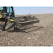 Side view of skid steer with rock reel attachment engaging loose soil and small rocks on dry field