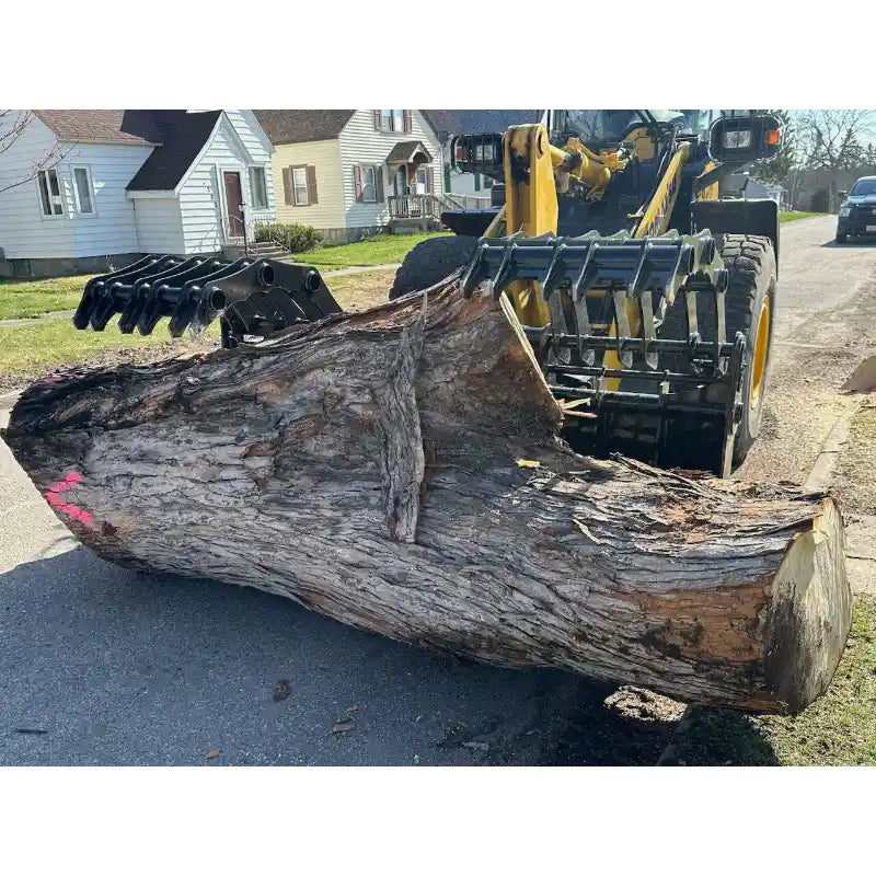 Yellow loader with black grapple attachment holding large cut tree trunk on residential street pavement