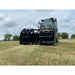 Front view of black steel clam-style grapple attachment mounted on loader over dry grass field under cloudy sky