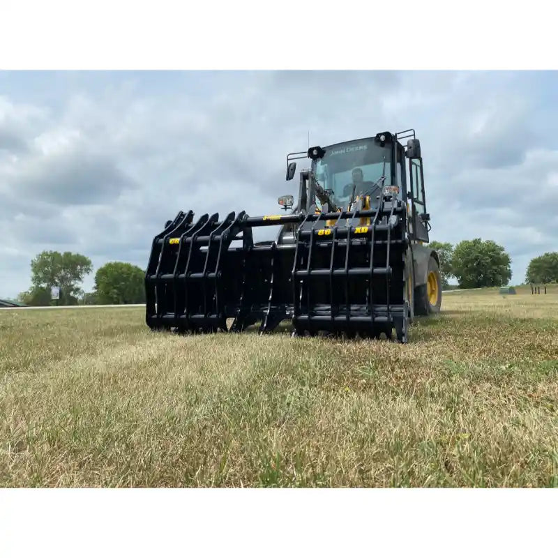 Front view of black steel clam-style grapple attachment mounted on loader over dry grass field under cloudy sky