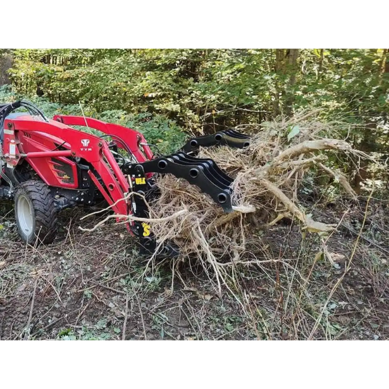 Red compact tractor with black grapple attachment lifting uprooted tree with exposed roots in wooded area